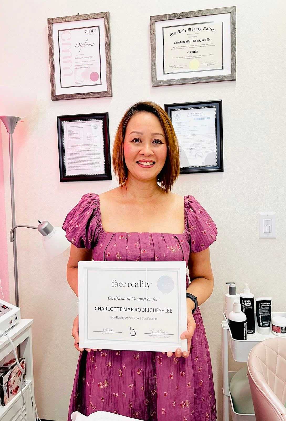 Woman in pink dress holding a certificate, standing in front of framed documents on a wall.